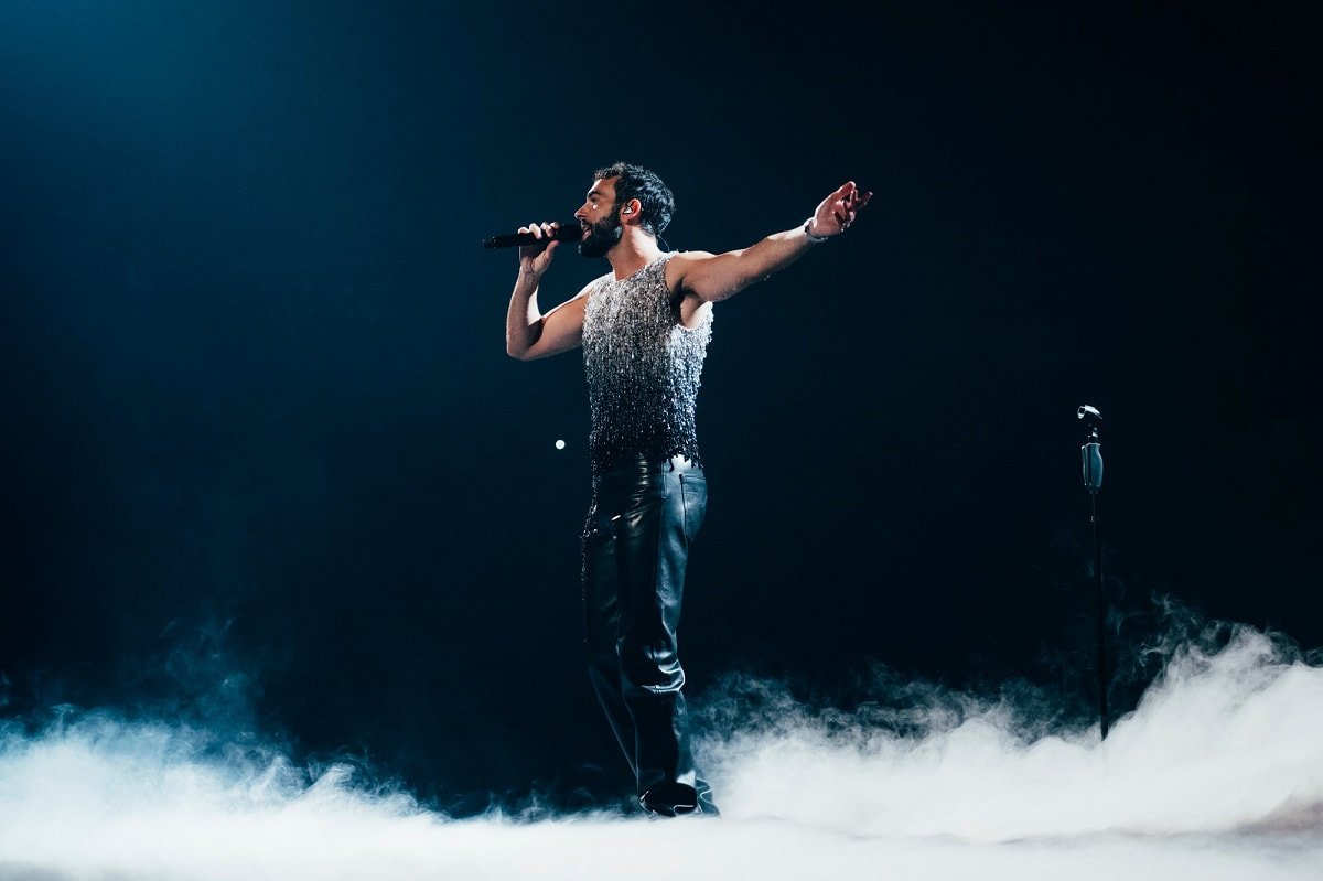 Man singing on stage with microphone wearing sleeveless top and leather pants, surrounded by smoke in a dark, dramatic setting.
