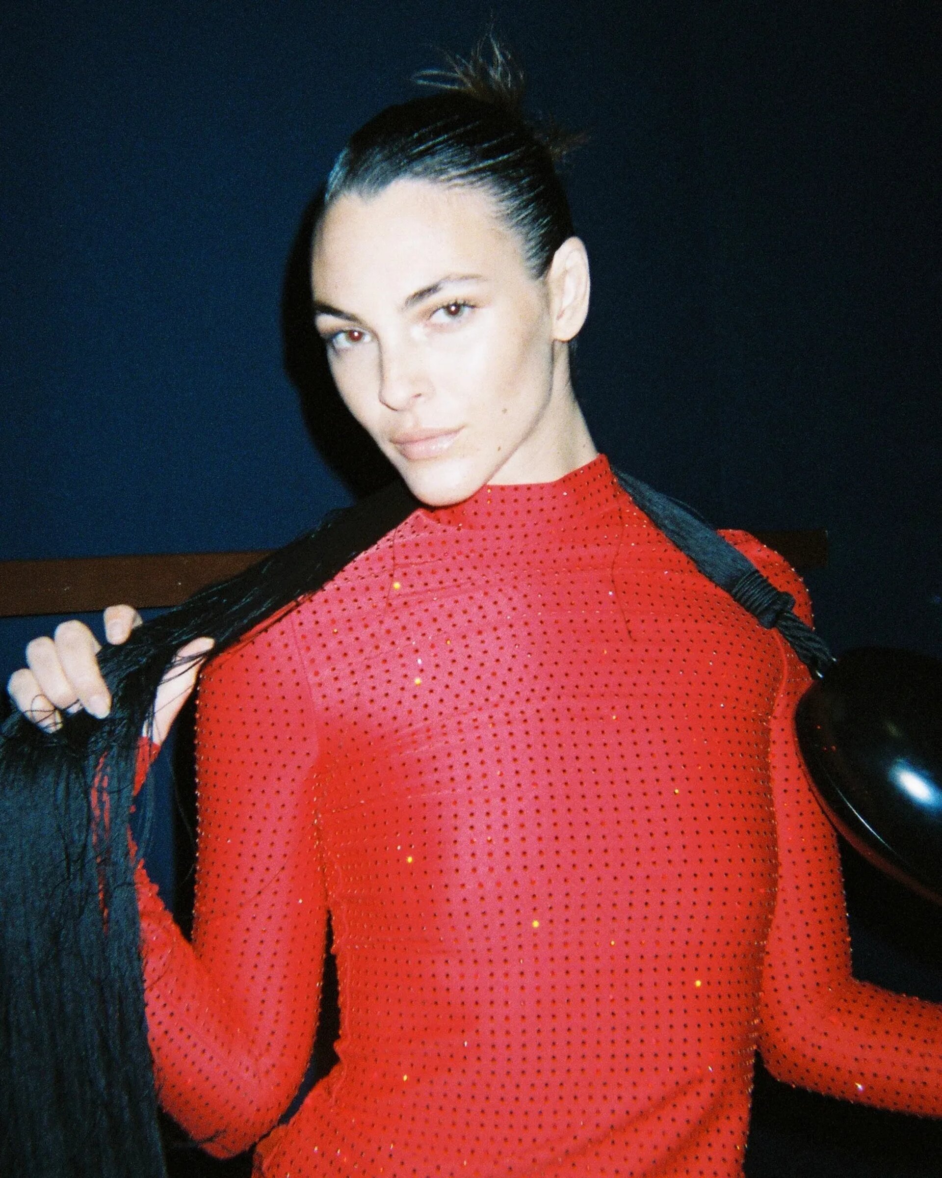 Portrait of a woman in a red, sparkly dress holding long black braids, indoors against a dark background with soft, diffused lighting.