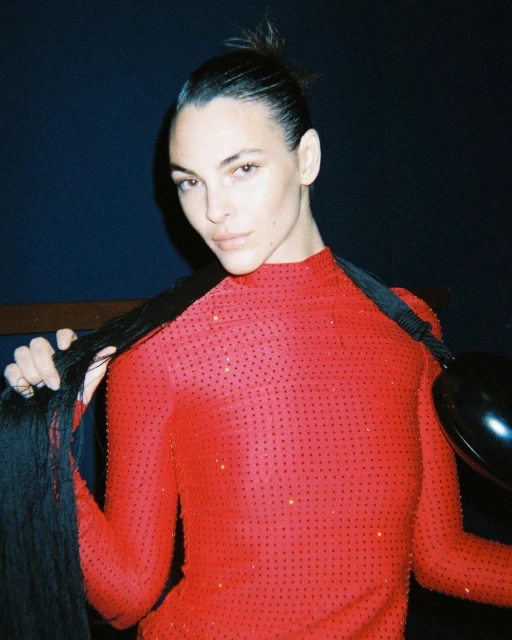 Portrait of a woman in a red, sparkly dress holding long black braids, indoors against a dark background with soft, diffused lighting.