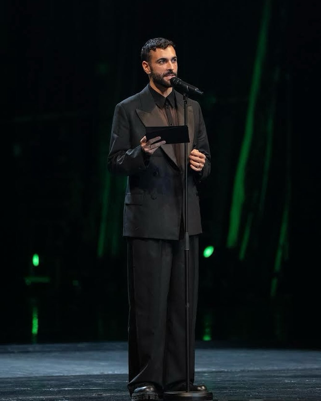 Man on stage giving a speech, wearing an all-black suit, holding a black object, with a microphone in front of a green-lit background.