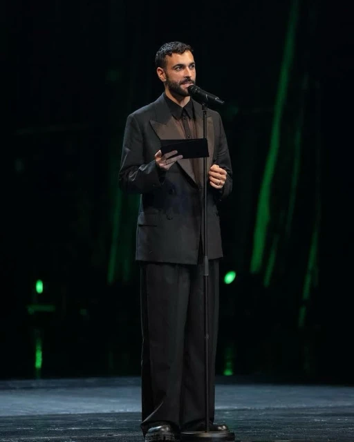 Man on stage giving a speech, wearing an all-black suit, holding a black object, with a microphone in front of a green-lit background.