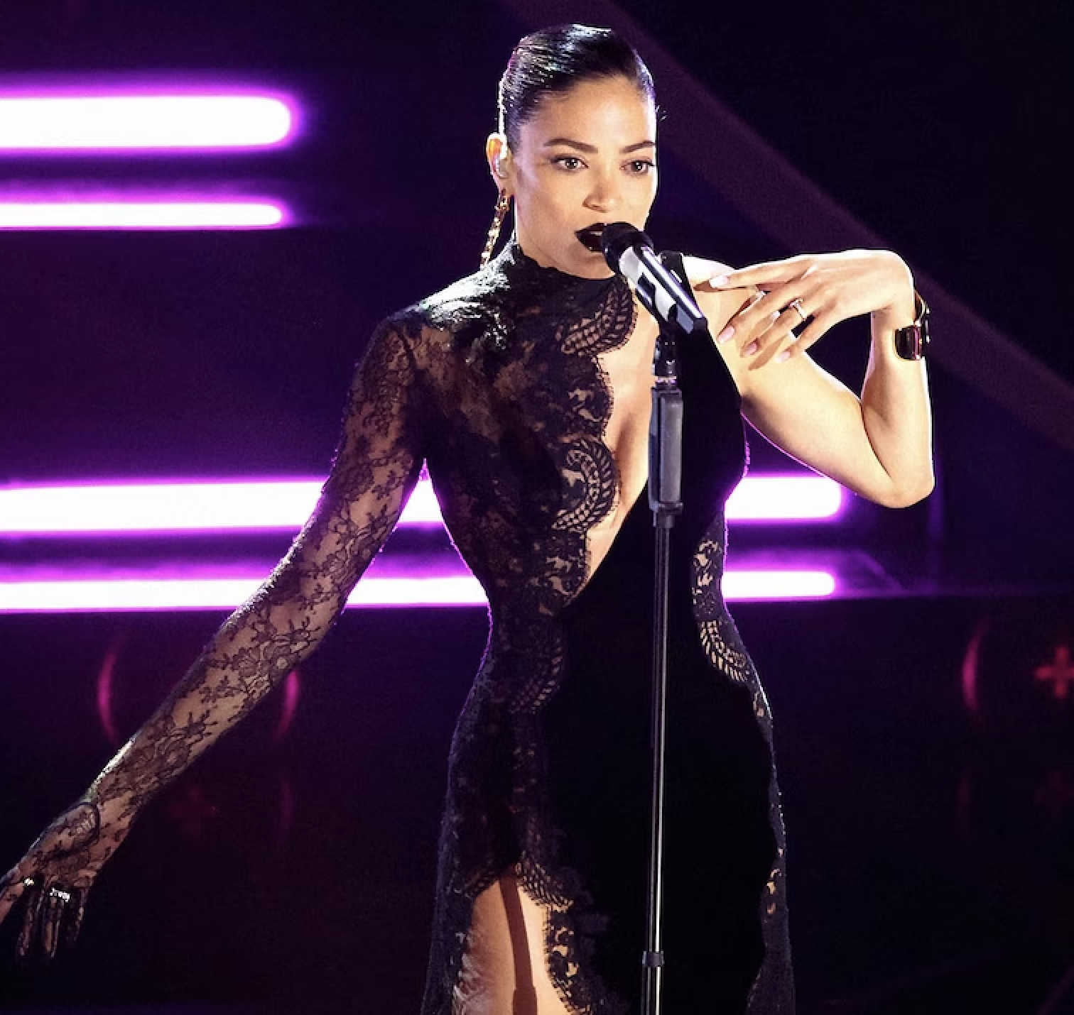 Singer in elegant black lace dress performs on stage with microphone. Purple and white lights illuminate the background, creating a vibrant atmosphere.