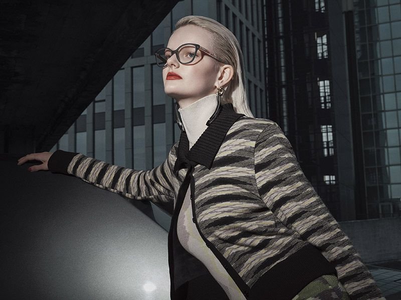 Stylish woman in patterned jacket and glasses poses against modern architecture for fashion editorial, showcasing sharp colors and details.