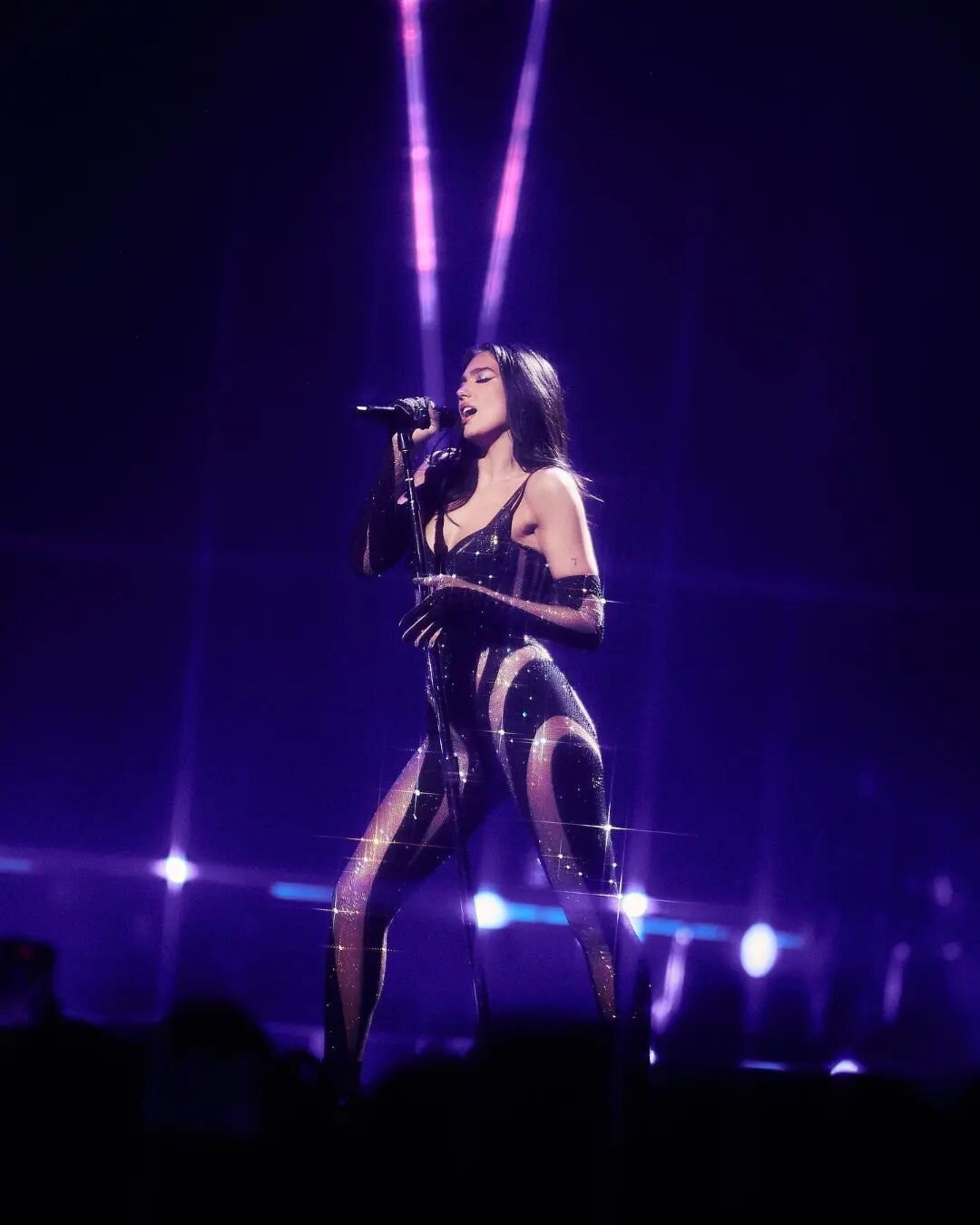 Dynamic stage shot of Dua Lipa singing, dressed in a sparkling black jumpsuit under violet stage lights, conveying energy and performance.