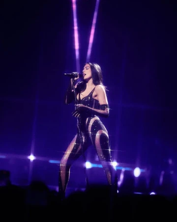Dynamic stage shot of Dua Lipa singing, dressed in a sparkling black jumpsuit under violet stage lights, conveying energy and performance.