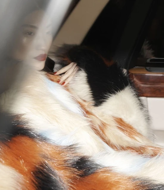 Portrait of an adult woman with red lipstick reclining in a car draped with a multicolor fur coat, posing in a relaxed manner.