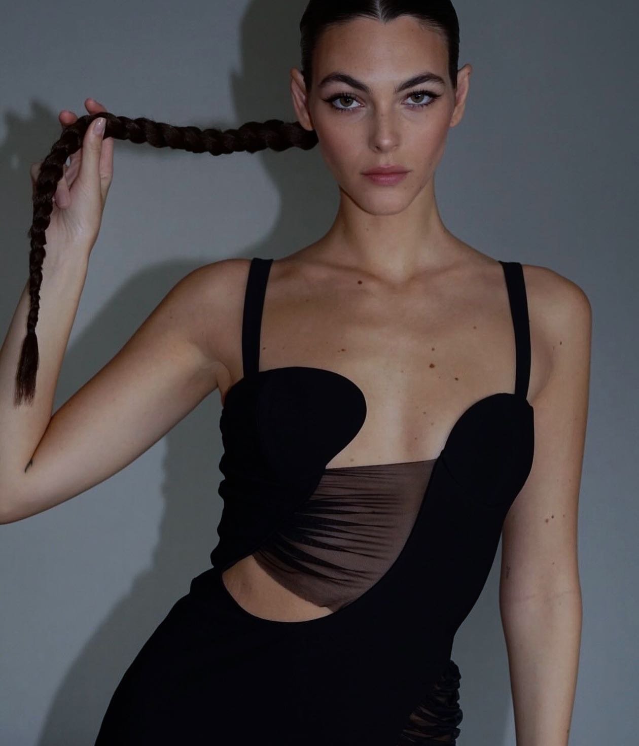 Brunette woman poses in a stylish black dress against a gray backdrop, holding a braid of her long hair up to her face.