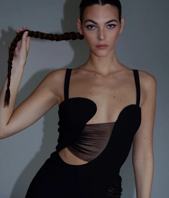 Brunette woman poses in a stylish black dress against a gray backdrop, holding a braid of her long hair up to her face.