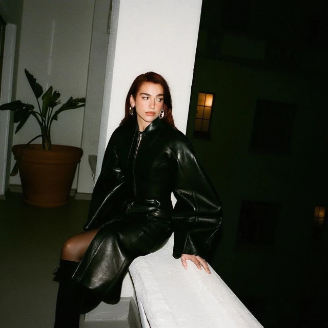 A woman with auburn hair sits on a ledge in a black leather dress and boots, with a potted plant in the background, during nighttime.