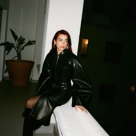 A woman with auburn hair sits on a ledge in a black leather dress and boots, with a potted plant in the background, during nighttime.