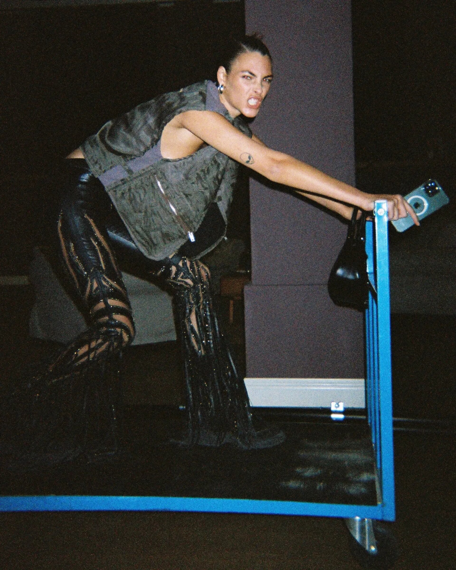 Fashionable adult female poses on a blue frame, wearing a vest, leather pants, and boots, holding a cell phone and small purse.