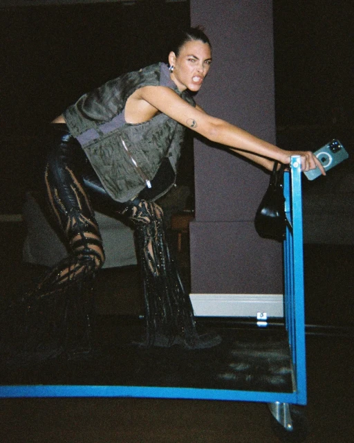 Fashionable adult female poses on a blue frame, wearing a vest, leather pants, and boots, holding a cell phone and small purse.