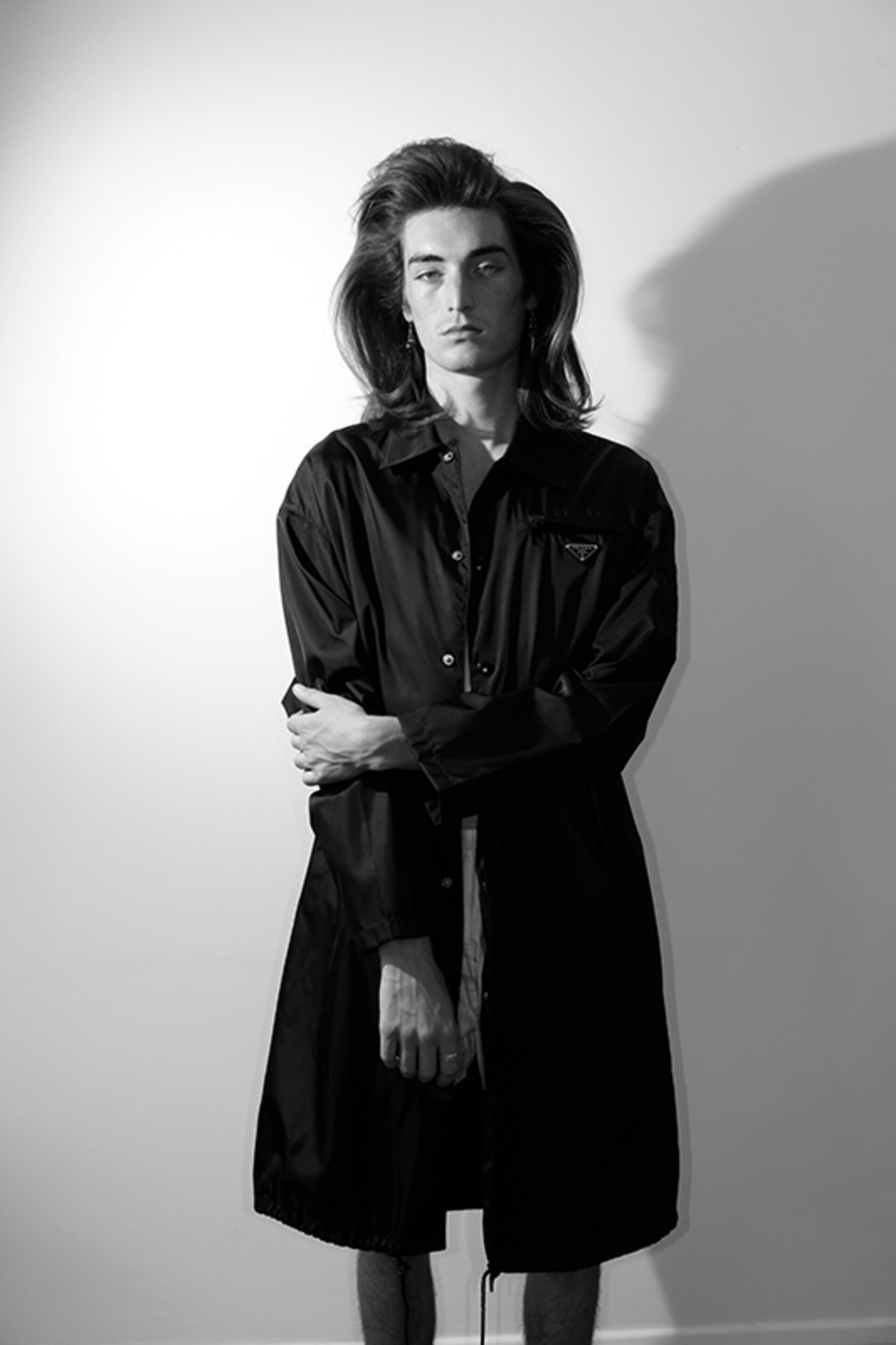 Portrait of a young male model with long hair wearing a black coat against a plain wall in Milan styled by Lorenzo Posocco.