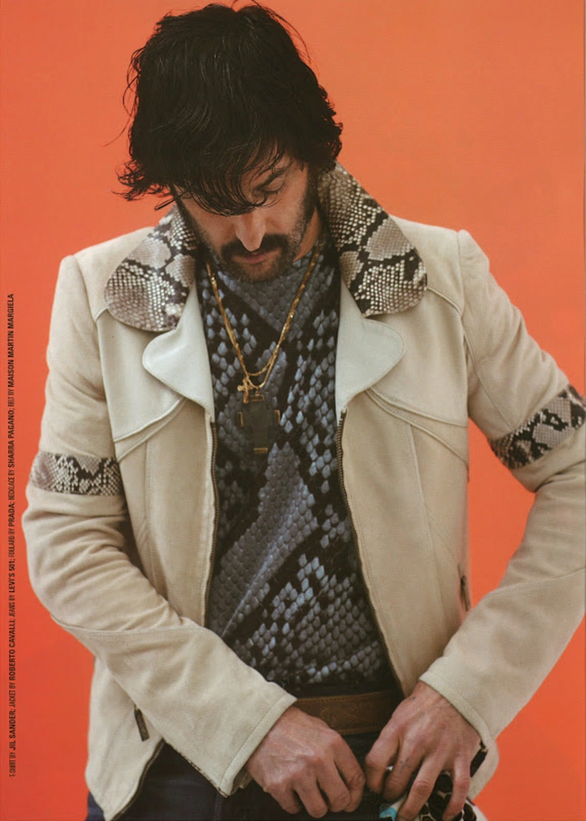 Man with dark hair and beard wearing an off-white jacket with snakeskin collar against an orange background