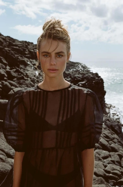 A young fair-skinned woman poses in a sheer, black dress against a background of rocky cliffs and ocean, captured in natural light.