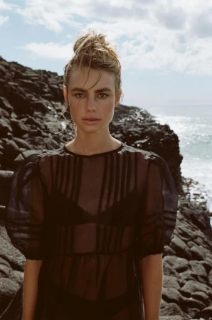 A young fair-skinned woman poses in a sheer, black dress against a background of rocky cliffs and ocean, captured in natural light.