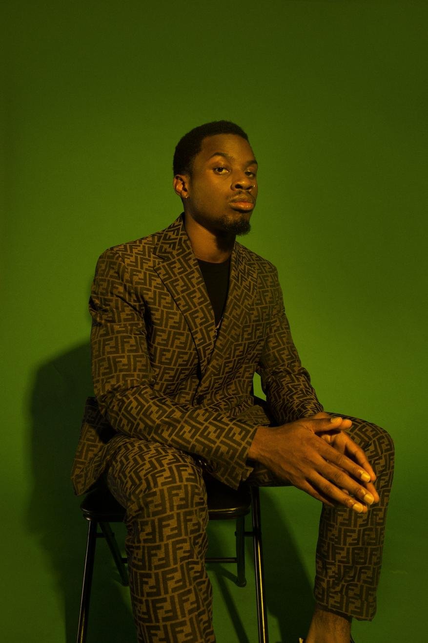 Portrait of an adult male sitting on a chair wearing a brown patterned suit in front of a green screen in a studio.