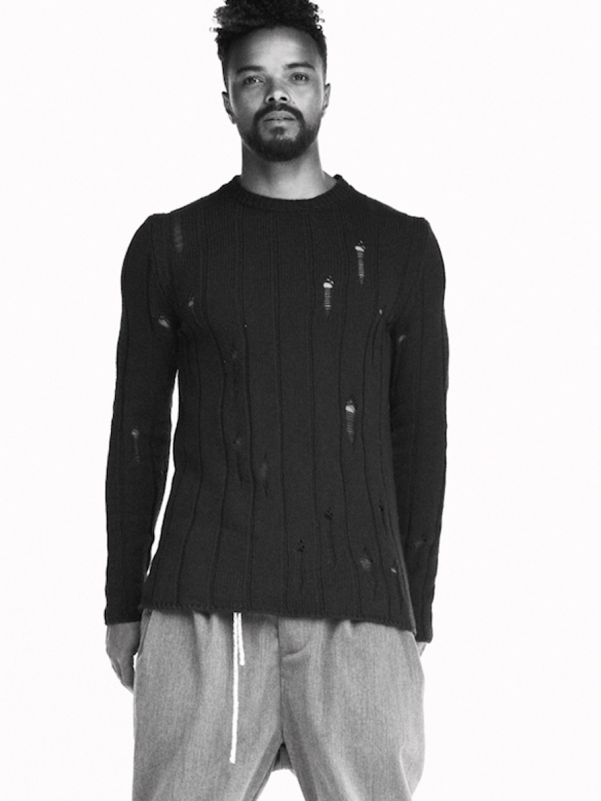 Adult male in black distressed sweater and grey pants against a white background in studio setting. Fashion advertisement photography.