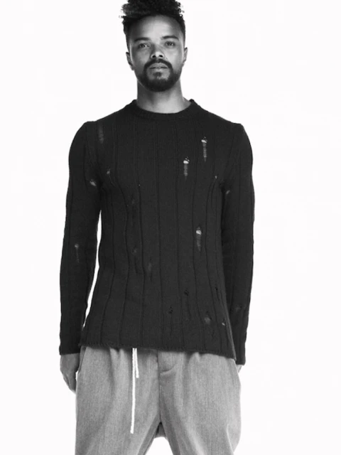 Adult male in black distressed sweater and grey pants against a white background in studio setting. Fashion advertisement photography.