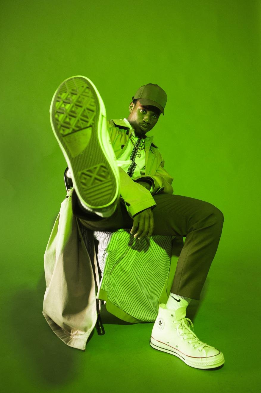 Man posing in green-toned studio with white sneakers, green cap, and trench coat. One leg extended with shoe toward camera.