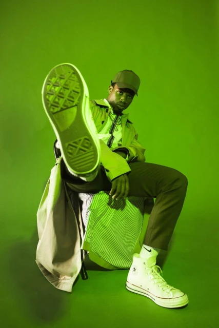 Man posing in green-toned studio with white sneakers, green cap, and trench coat. One leg extended with shoe toward camera.