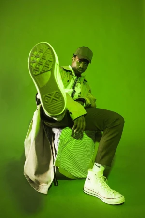 Man posing in green-toned studio with white sneakers, green cap, and trench coat. One leg extended with shoe toward camera.