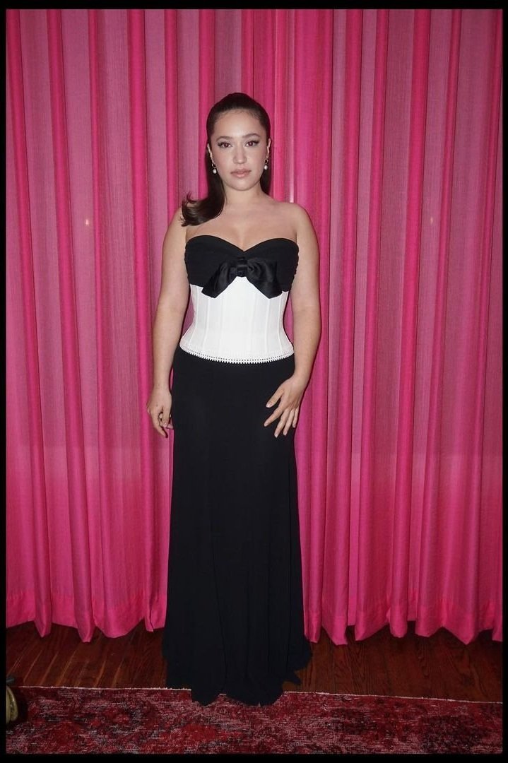 Portrait of a woman in formal evening attire. She is standing against a pink curtain backdrop with a red patterned rug on the floor.