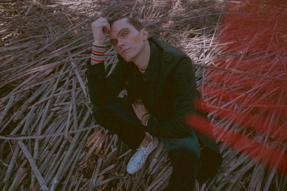 Adult man in a dark jacket poses while crouching on reeds. White sneakers and a ring on his finger complete the outdoor portrait.