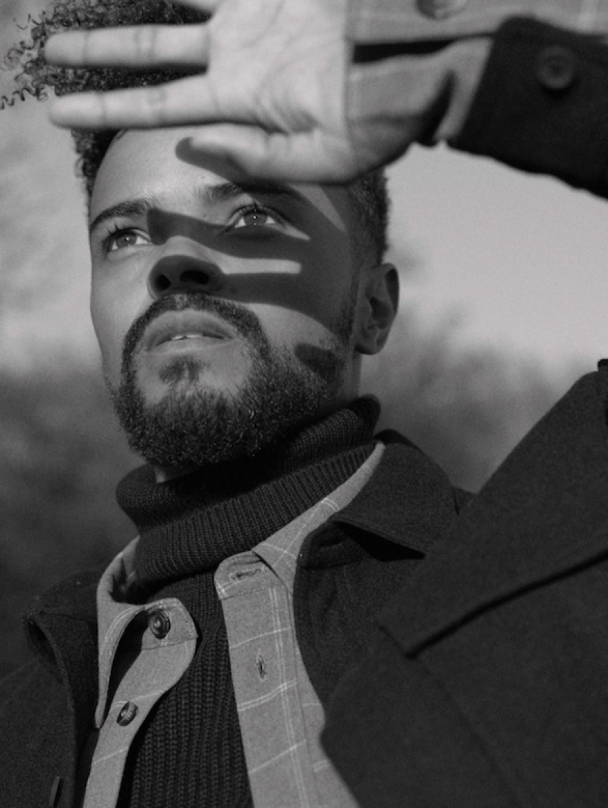 Monochrome portrait of a man looking up, holding his hand up to block bright light casting shadows across his face.