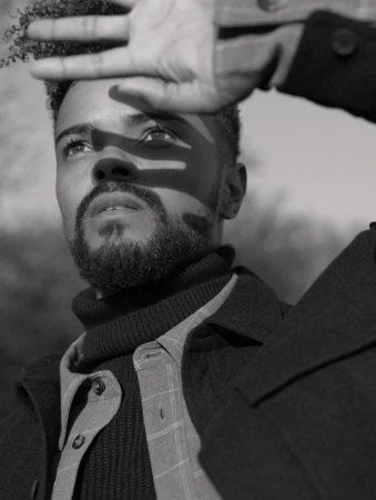 Monochrome portrait of a man looking up, holding his hand up to block bright light casting shadows across his face.