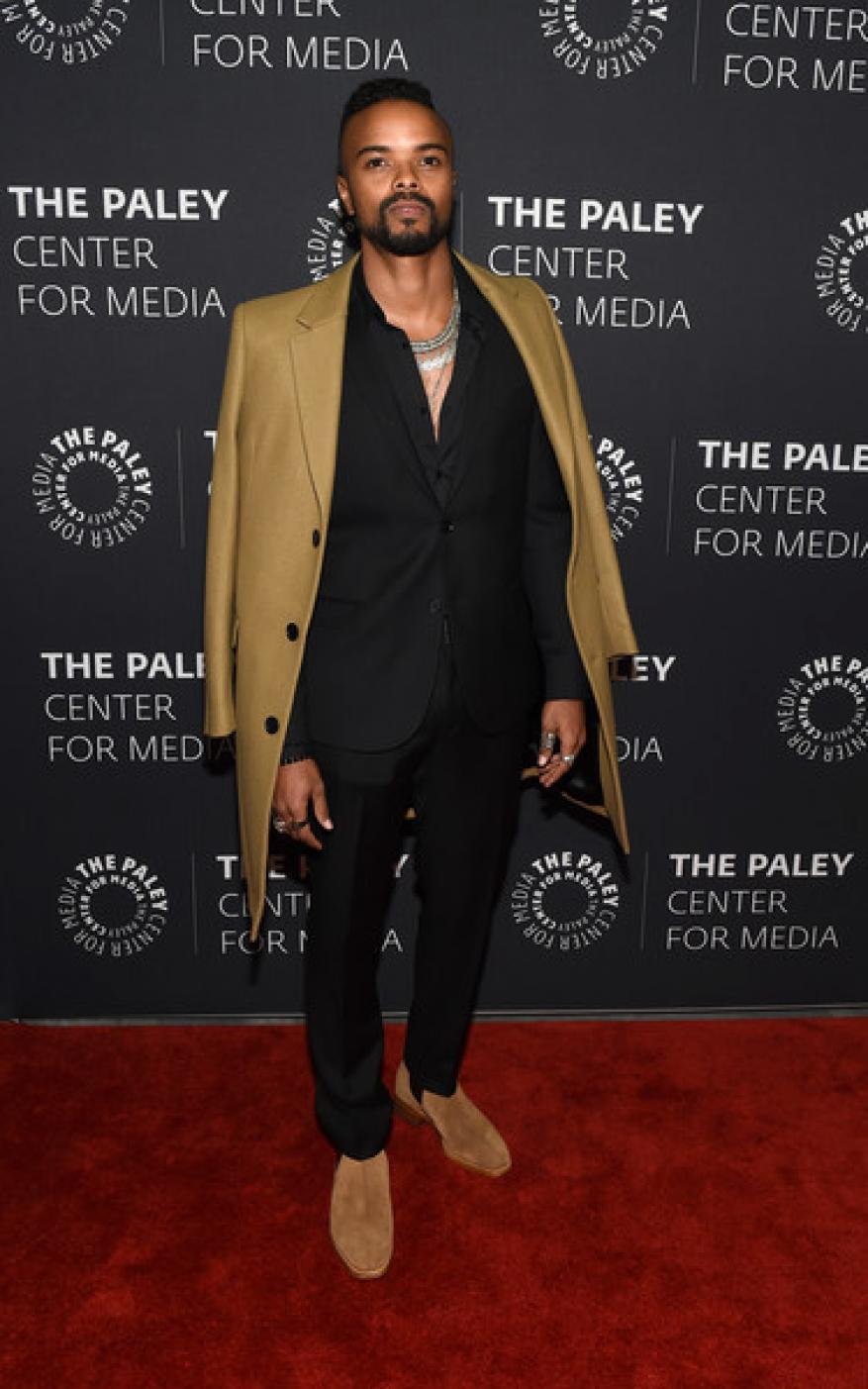 A full shot of a Black man in stylish formalwear and a tan coat draped over his shoulder, posing confidently against a branded backdrop.