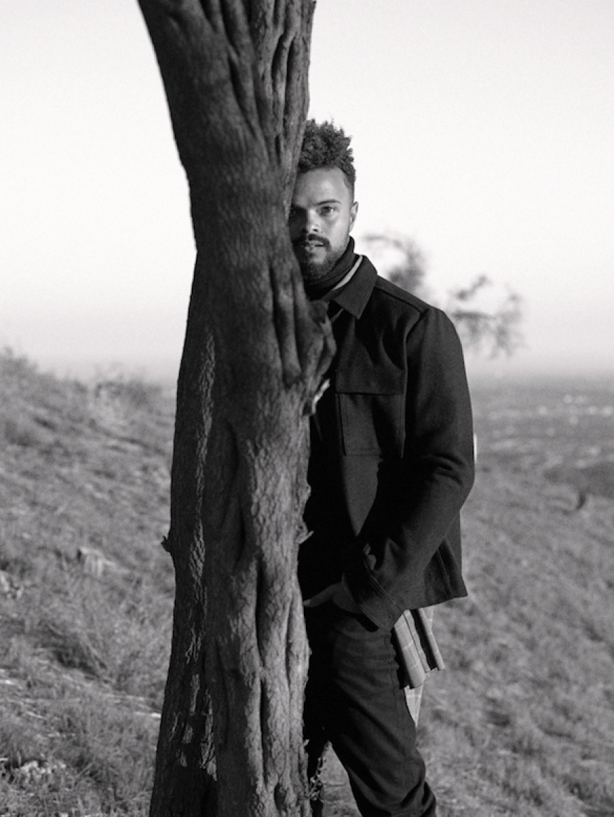 Monochrome portrait of a man peeking from behind a tree trunk on a hillside, styled by Lucy Warren in Los Angeles.