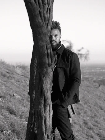 Monochrome portrait of a man peeking from behind a tree trunk on a hillside, styled by Lucy Warren in Los Angeles.