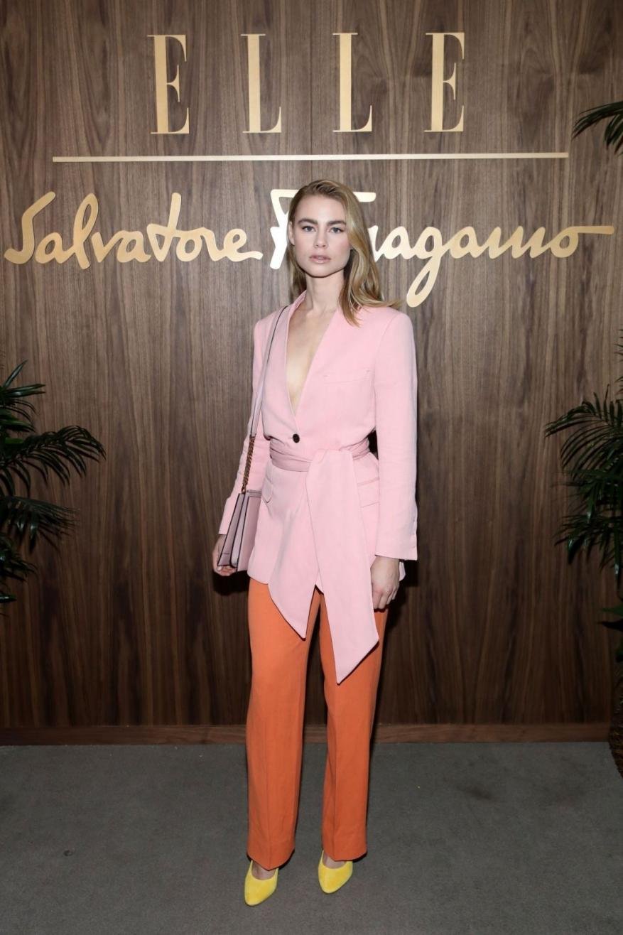 A stylish woman in a pink blazer and orange pants poses in front of the ELLE and Salvatore Ferragamo logos on a wood-paneled wall.