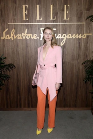 A stylish woman in a pink blazer and orange pants poses in front of the ELLE and Salvatore Ferragamo logos on a wood-paneled wall.