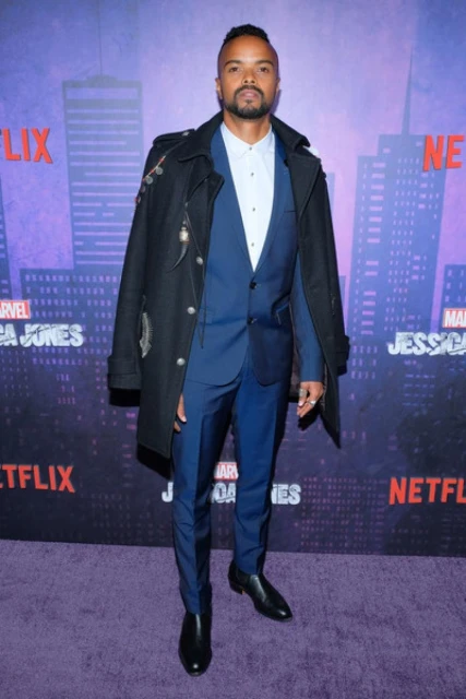 Man in a blue suit with a black coat on a purple carpet at an event. The backdrop is a city skyline with event branding.