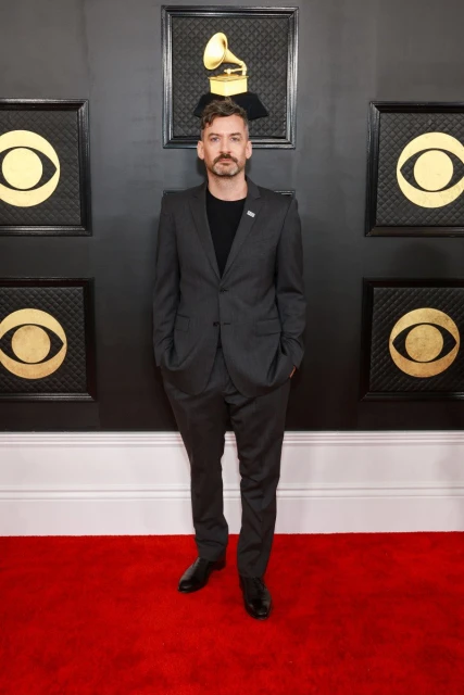 A man poses on a red carpet in a dark suit jacket and pants in front of Grammy-themed decoration at a music award show.