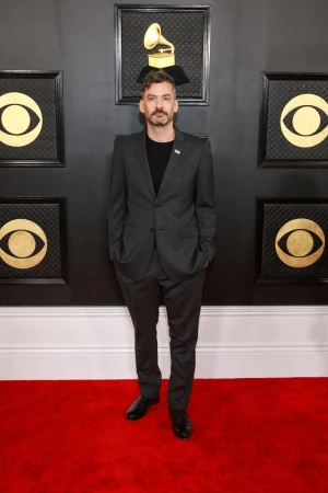 A man poses on a red carpet in a dark suit jacket and pants in front of Grammy-themed decoration at a music award show.