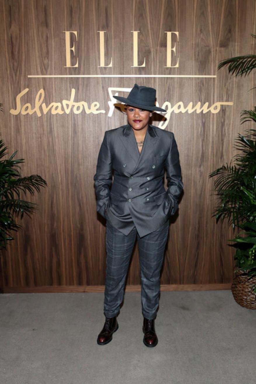A person in a grey suit and hat poses in front of an ELLE and Salvatore Ferragamo branded backdrop with potted plants.