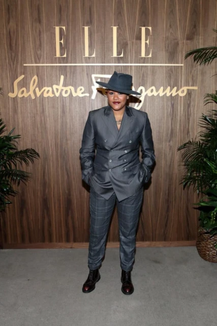 A person in a grey suit and hat poses in front of an ELLE and Salvatore Ferragamo branded backdrop with potted plants.