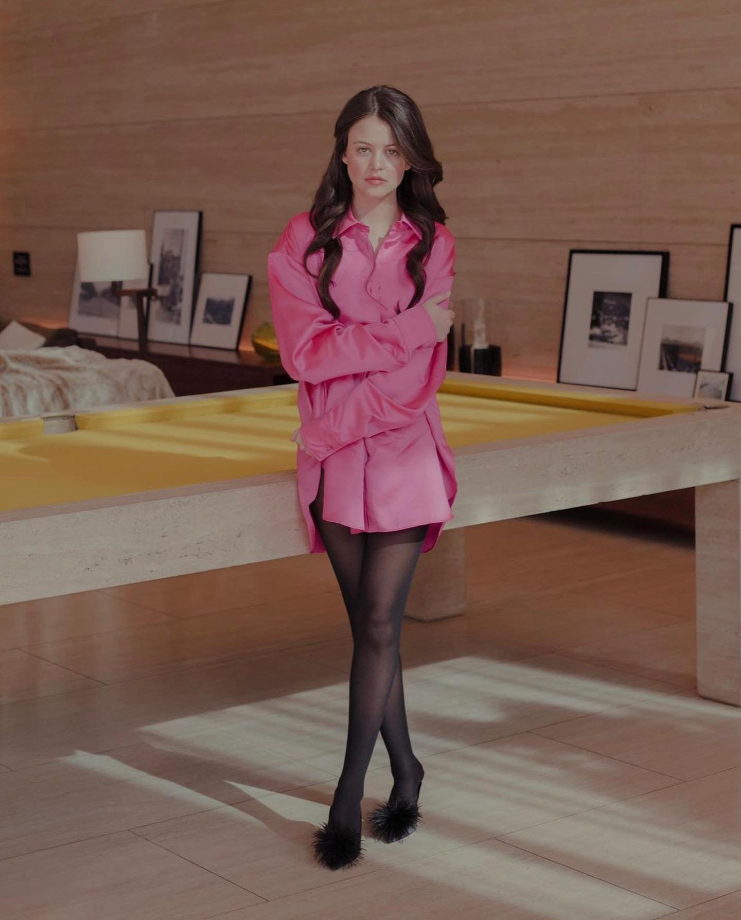 A brunette woman poses indoors wearing an oversized pink shirt, matching shorts, sheer tights, and black, feathered, open-toe high heels.