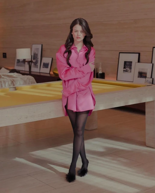 A brunette woman poses indoors wearing an oversized pink shirt, matching shorts, sheer tights, and black, feathered, open-toe high heels.