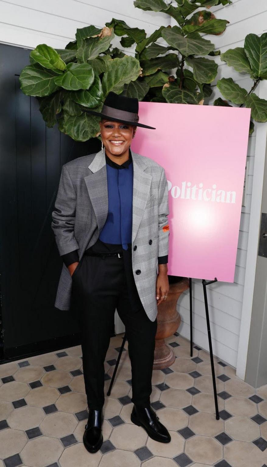 Adult female in an oversized blazer and hat poses next to a 'Politician' poster in a bright indoor setting with greenery visible.