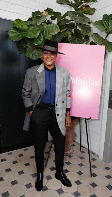 Adult female in an oversized blazer and hat poses next to a 'Politician' poster in a bright indoor setting with greenery visible.