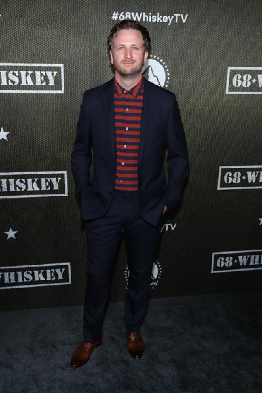 Man in a suit standing against a backdrop with a show logo. He has curly blond hair and is wearing a striped shirt and dress shoes.