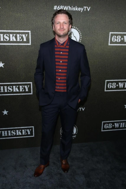 Man in a suit standing against a backdrop with a show logo. He has curly blond hair and is wearing a striped shirt and dress shoes.