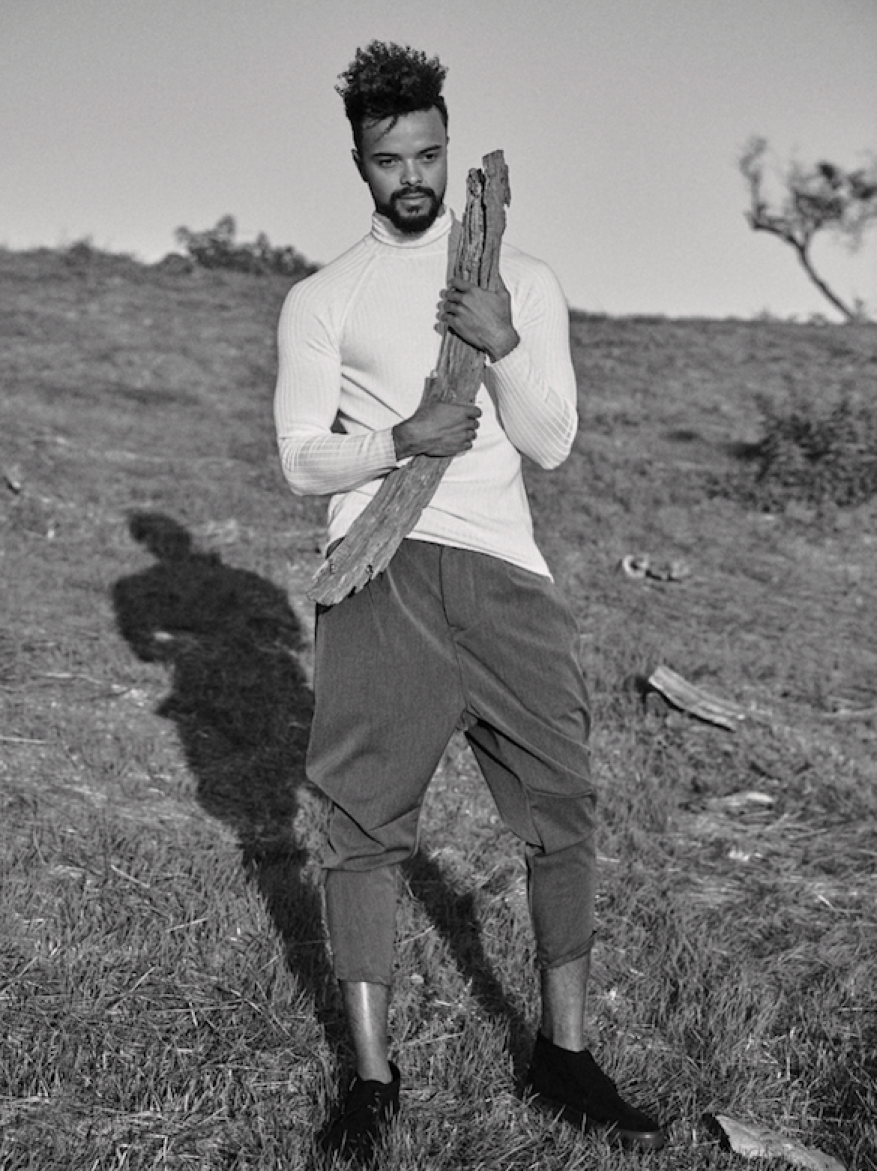 Monochrome portrait of a man standing in a grassy field holding a weathered piece of wood, wearing a long-sleeved shirt and cropped pants.