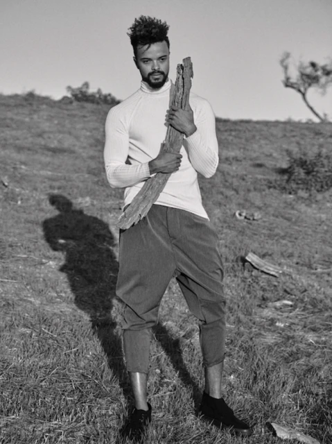 Monochrome portrait of a man standing in a grassy field holding a weathered piece of wood, wearing a long-sleeved shirt and cropped pants.