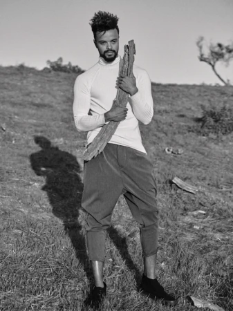 Monochrome portrait of a man standing in a grassy field holding a weathered piece of wood, wearing a long-sleeved shirt and cropped pants.