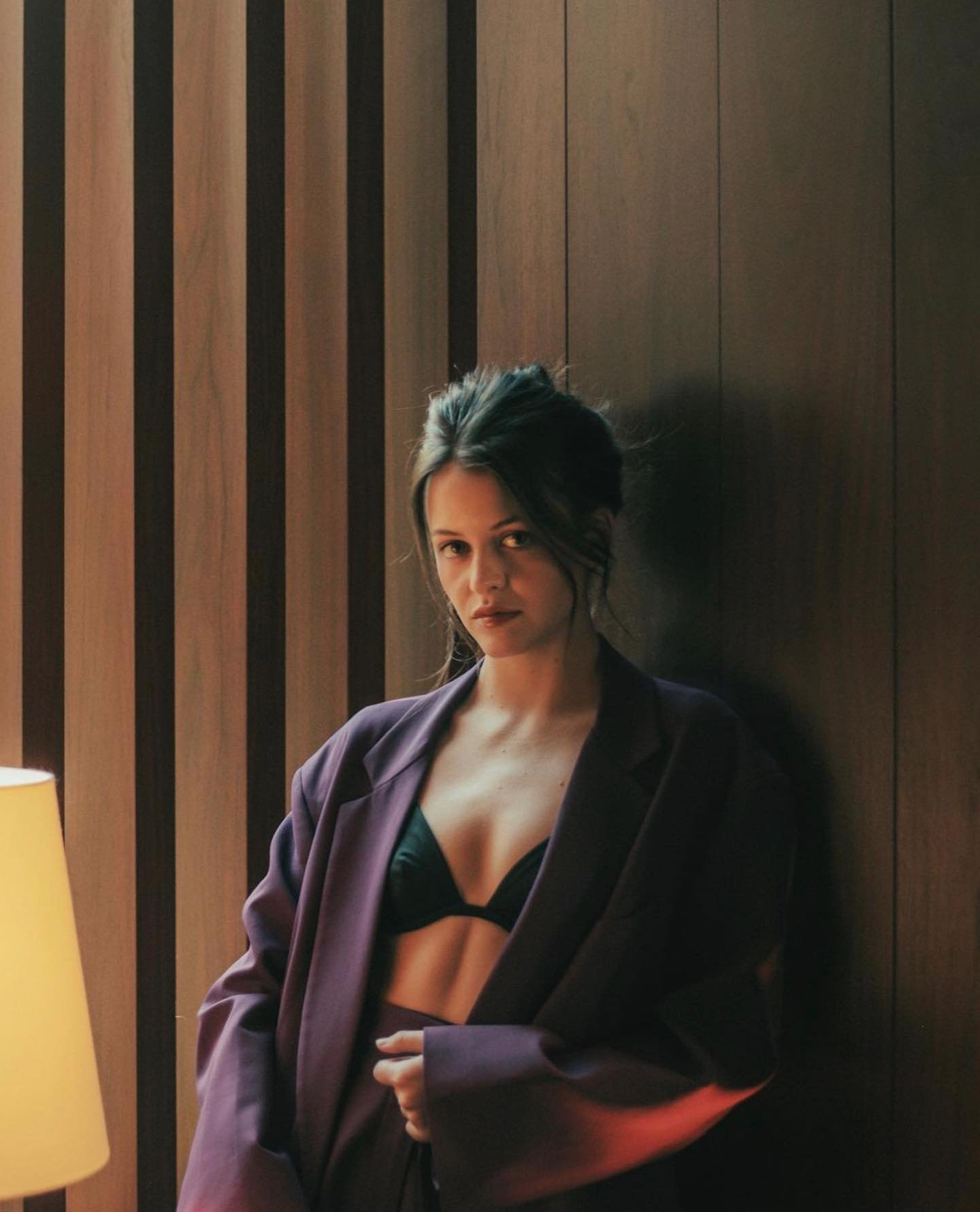 Attractive woman in oversized purple blazer and black bra poses indoors against a striped wood wall, with a table lamp visible.